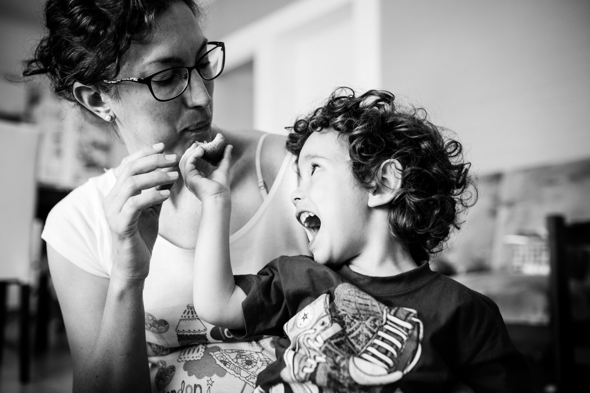 Toddler feeding his mom, giving her a strawberry while opening his mouth very big. Black and white photography.