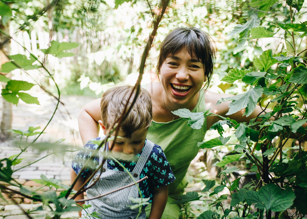 Woman with a very big smile on her face, she's playing with his son in their backyard. There's a lot of green on the image. Documentary family photographer amsterdam