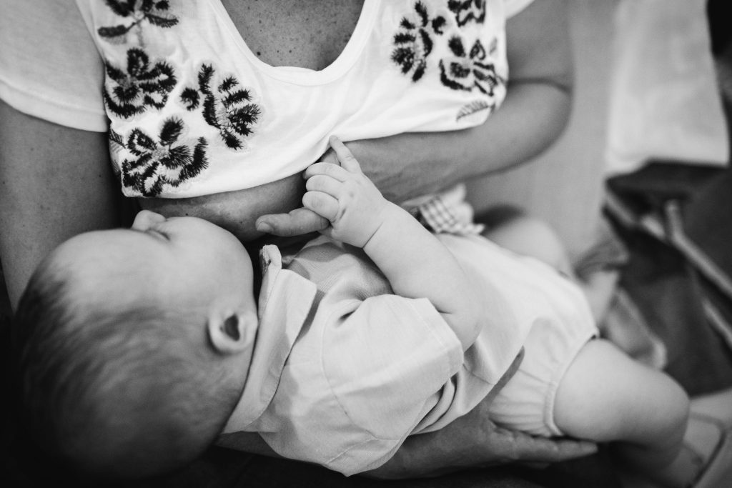 Mom breastfeeding her six month old baby in their home, while baby holds her finger in a documentary family photoshoot