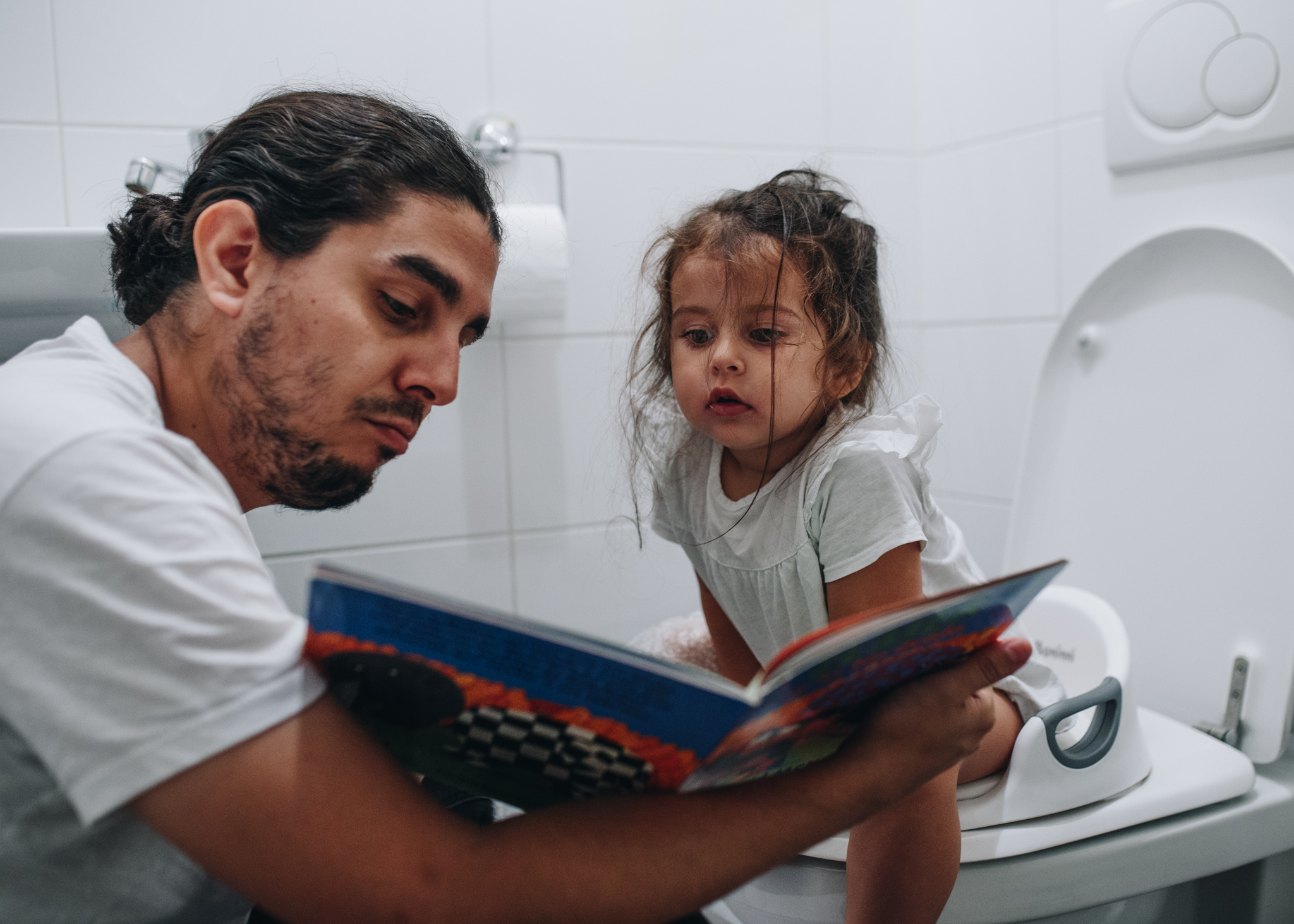 Dad reading a book to his daughter while she's sitting on the toilet