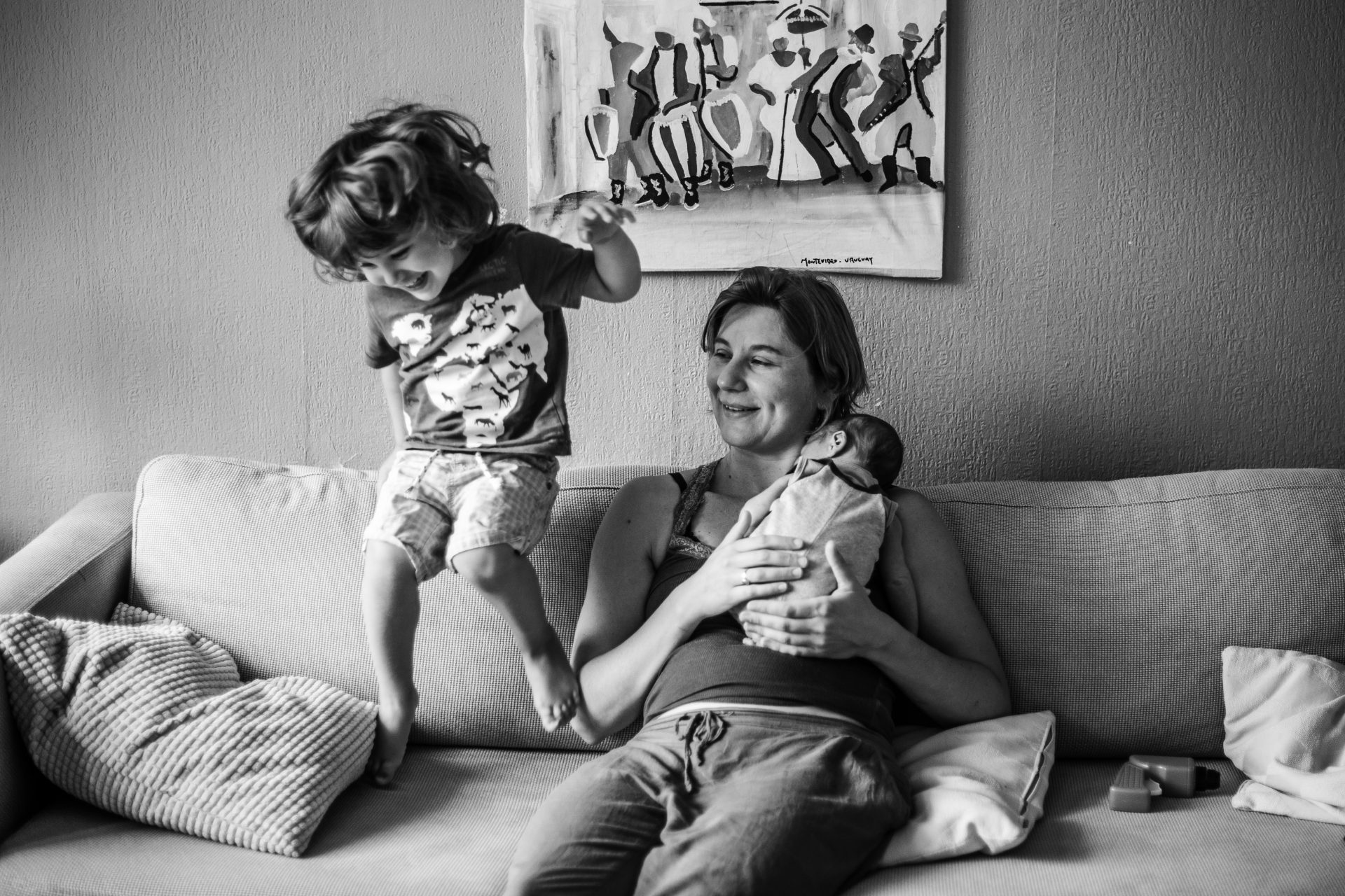 Mom holding her newborn in her chest sitting on the couch while her toddler is jumping on it next to her. Documentary photoshoot in Amsterdam.