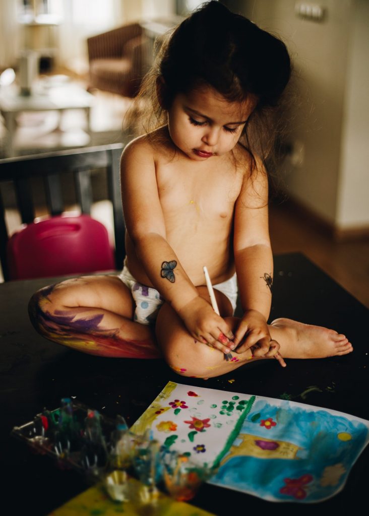 Toddler girl on the table painting her own legs with temperas. Natural style photographer. Amsterdam.