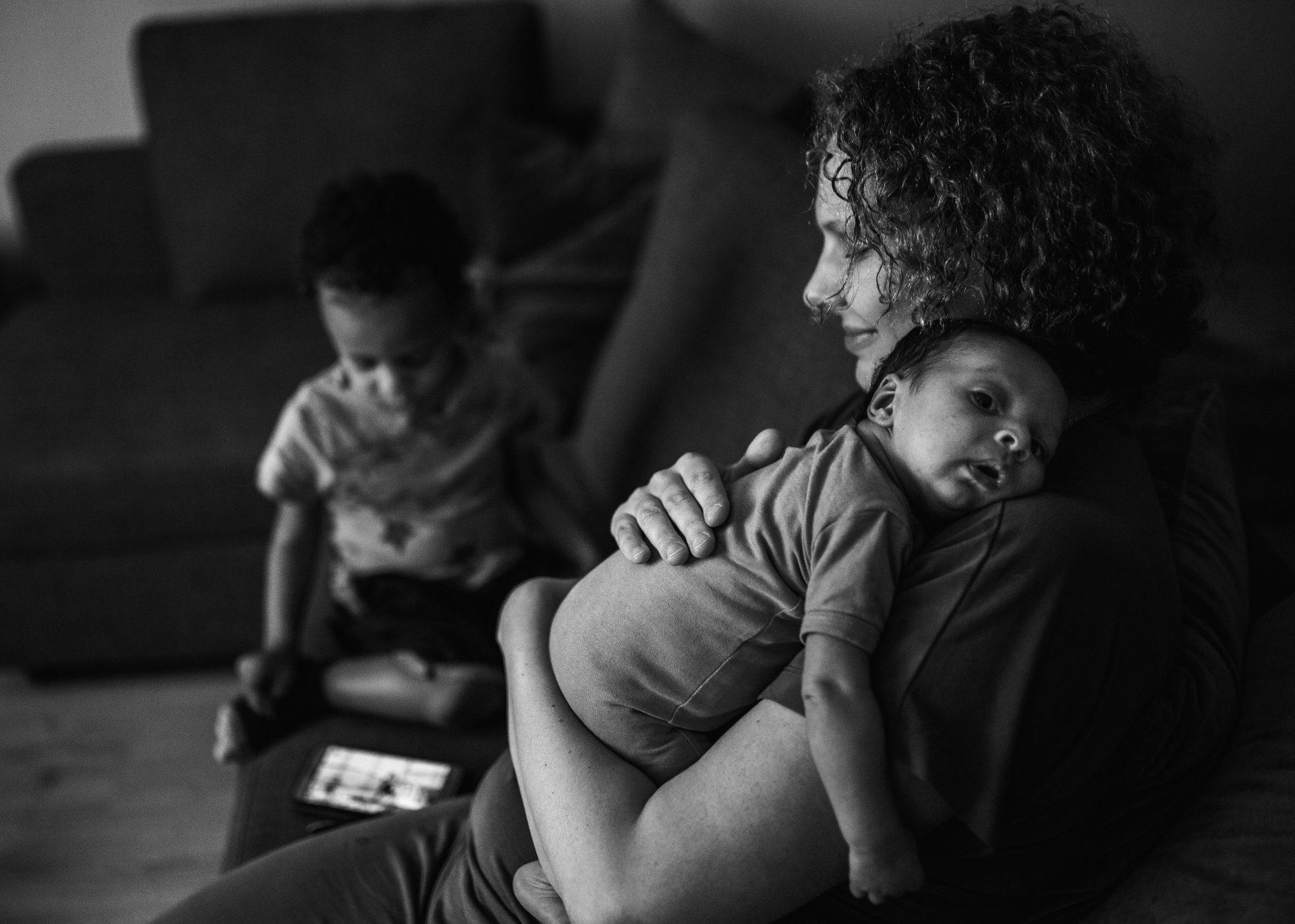 Black and white picture of mom holding her newborn in the livingroom with a calm smile, with her toddler next to her playing with a cellphone