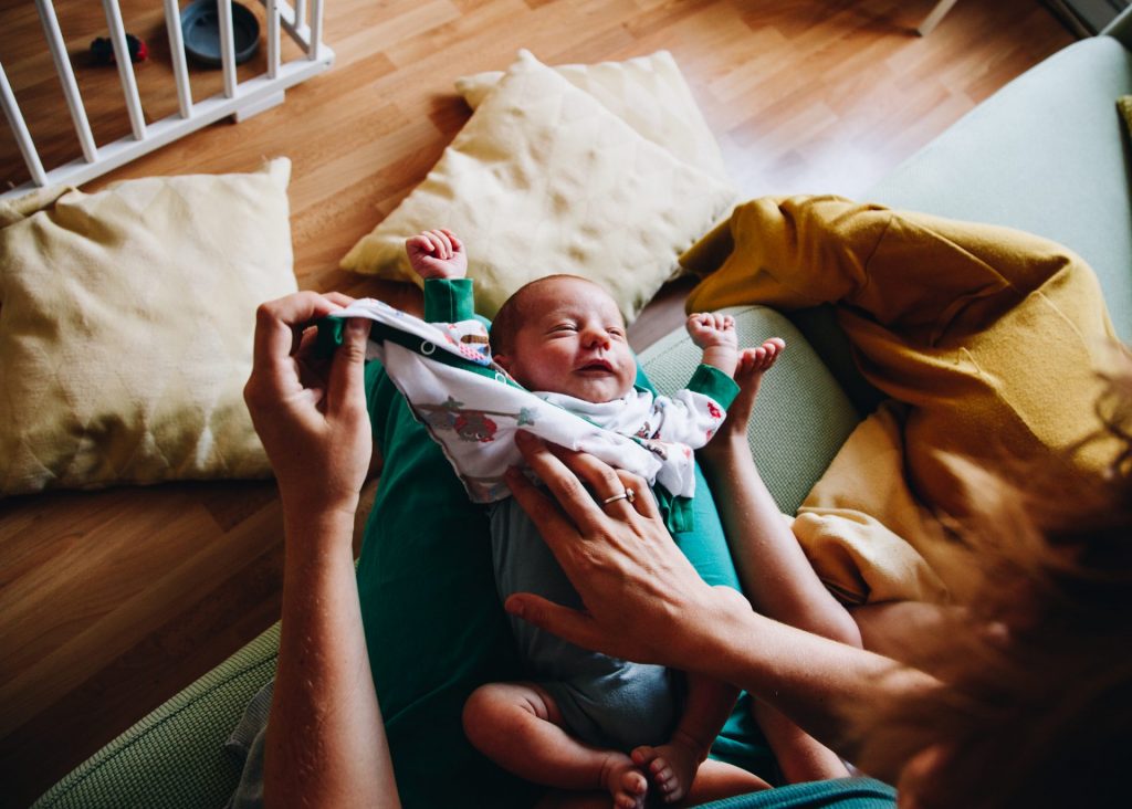 Newborn stretching his arms while his mom is gently changing his clothes. He lies on his mom's lap. Bird view angle. They're on the couch, sitting next to his older sister.