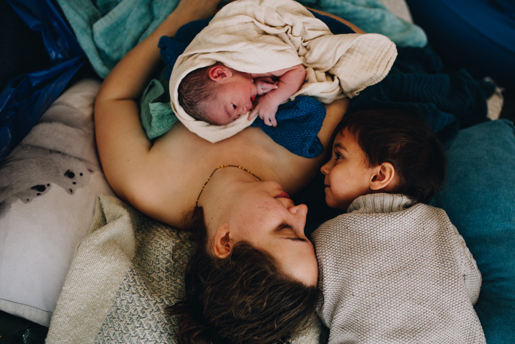 Geboorte fotografie. After giving birth to her second child, mom is lying on the bad, holding her newborn and contemplating her older child. Amsterdam birth photography.