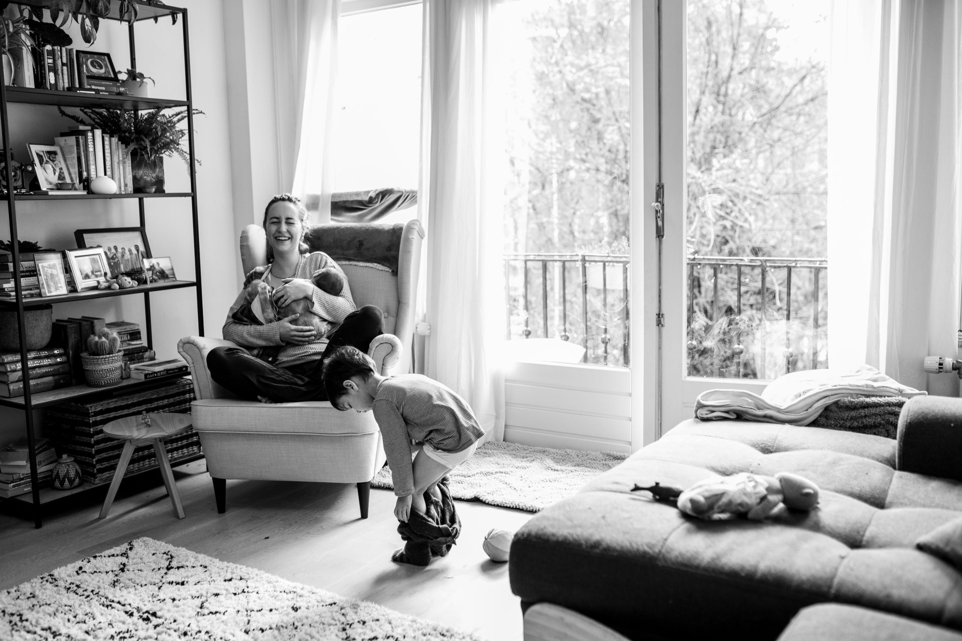 Mom holding her newborn while laughing hard because her older child is taking his own pants off. Black and white photo. In-home documentary session in Amsterdam.