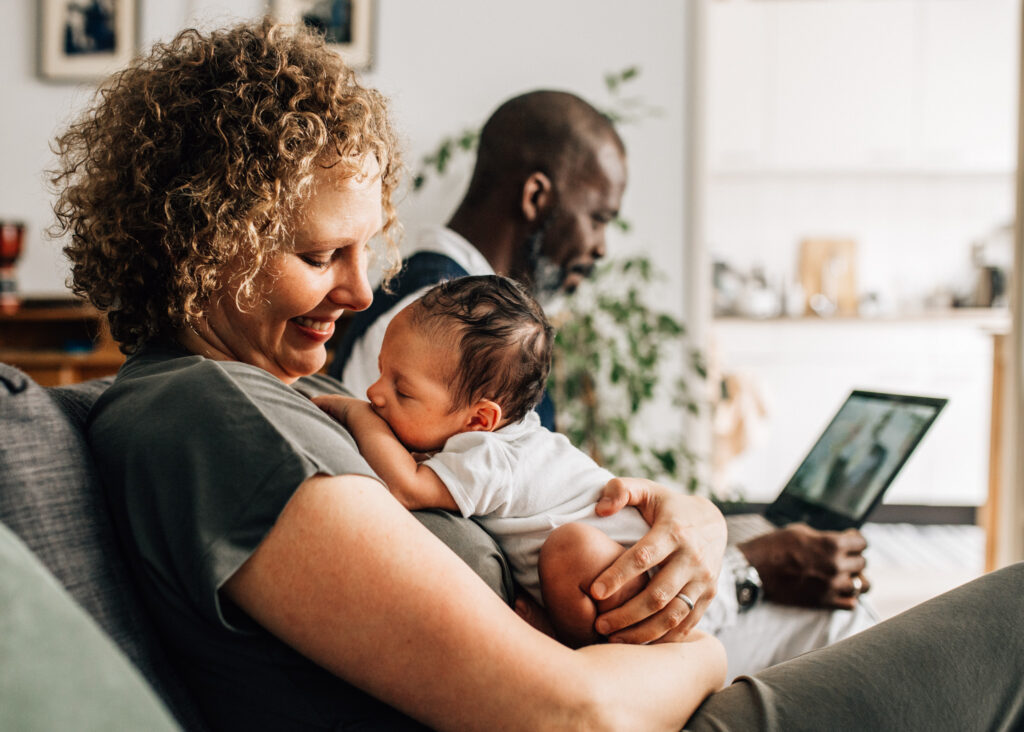 newborn nestling on mom's chest. She's looking at him happy, with a smile. Dad is behind them working on his computer. Amsterdam, Netherlands.