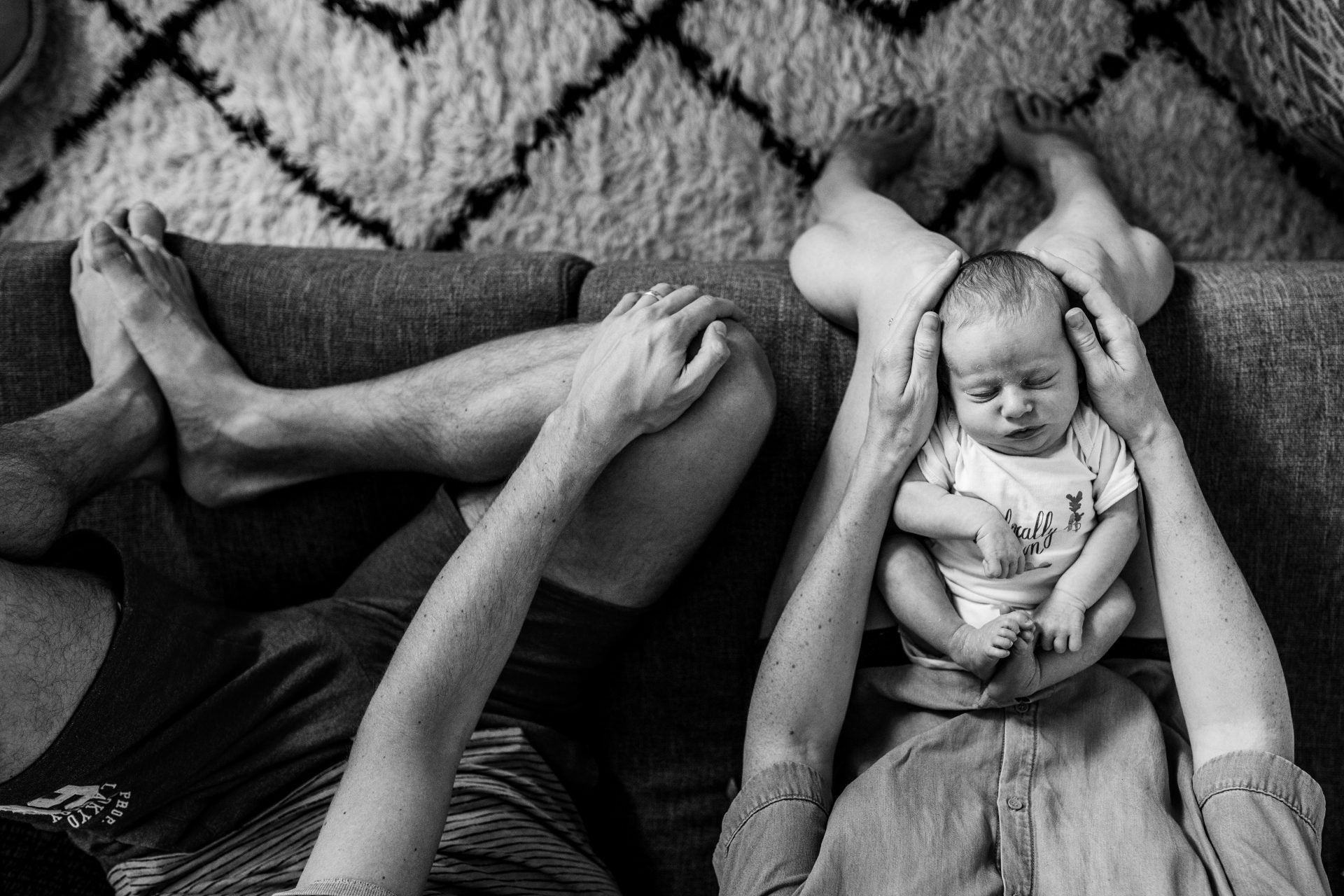 black and white photo of a newborn girl sleeping on her mom's lap. Her mom is gently holding her head. Next to them is her father. Bird view angle.