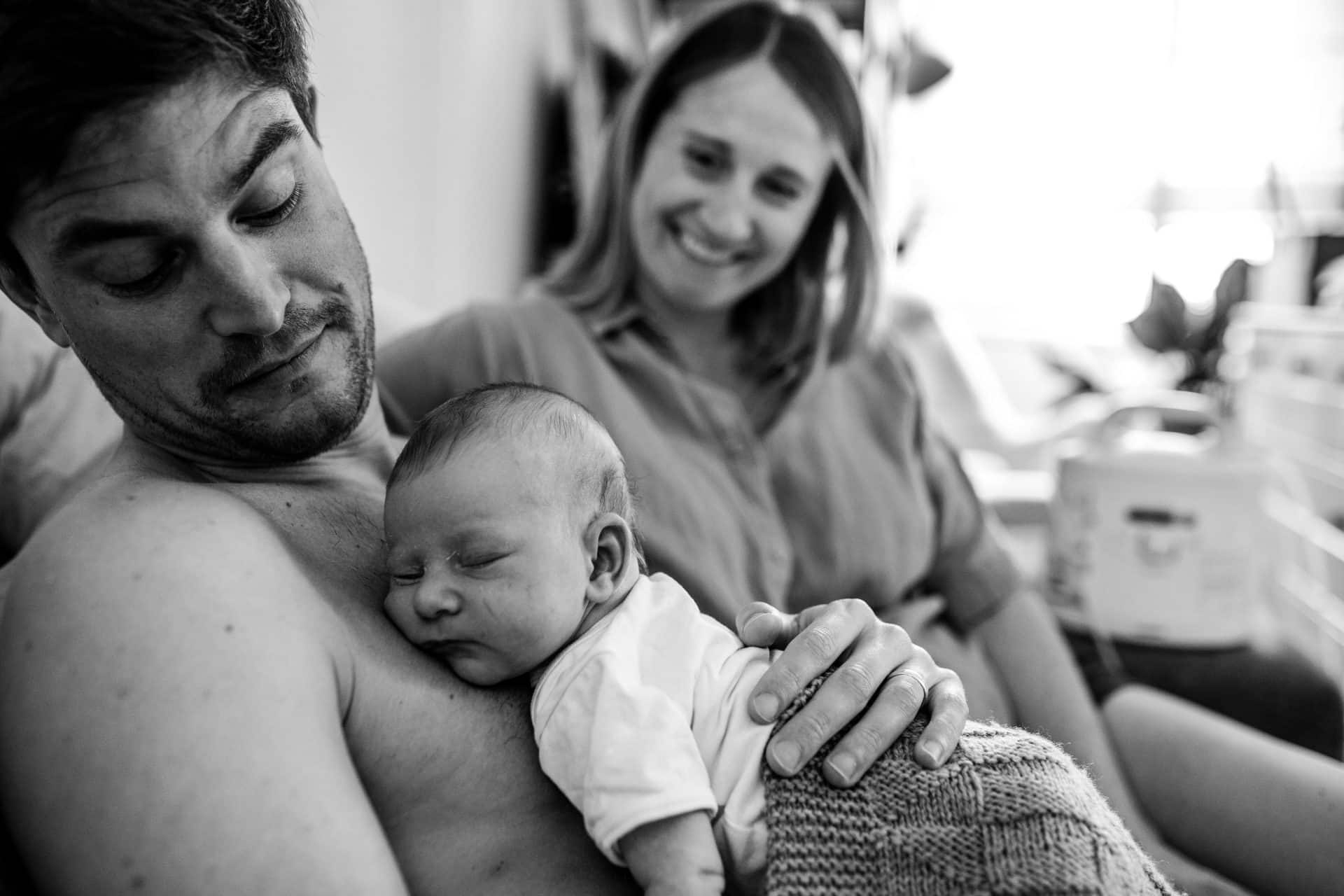 newborn sleeping on her father's chest, while he and her mom are contemplating at her