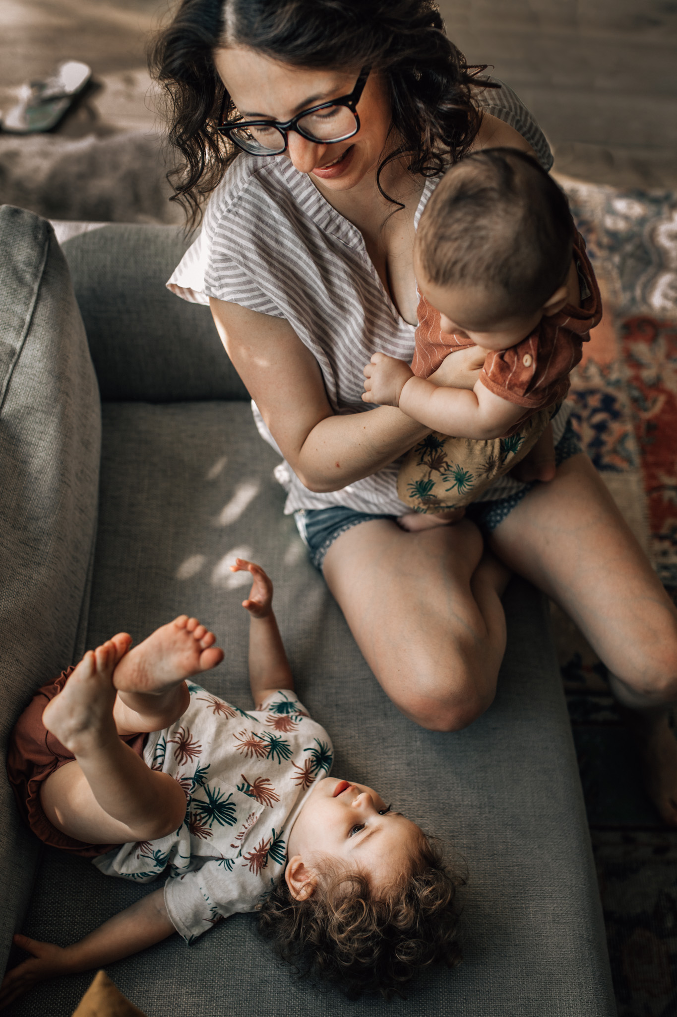 Family photography in Amsterdam: in the picture a mother with her two children is on the couch. She's holding her baby while her toddler is laying on the couch with her legs and feet pointing to the ceiling. The picture is taken from above.