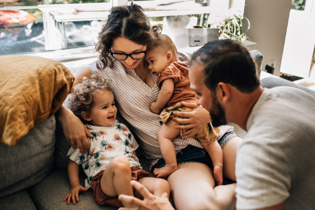 Family playing on the couch. Mom is holding her baby boy on her chest and next to them, her little toddler is sitting, smiling. Dad is playing to chase the little one. They're wearing soft, light, earthy tones with soft patterns.