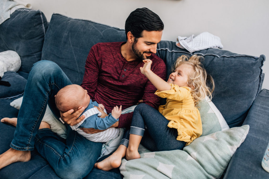 Portrait of a father with his newborn and older kid playing on the couch of their house. 