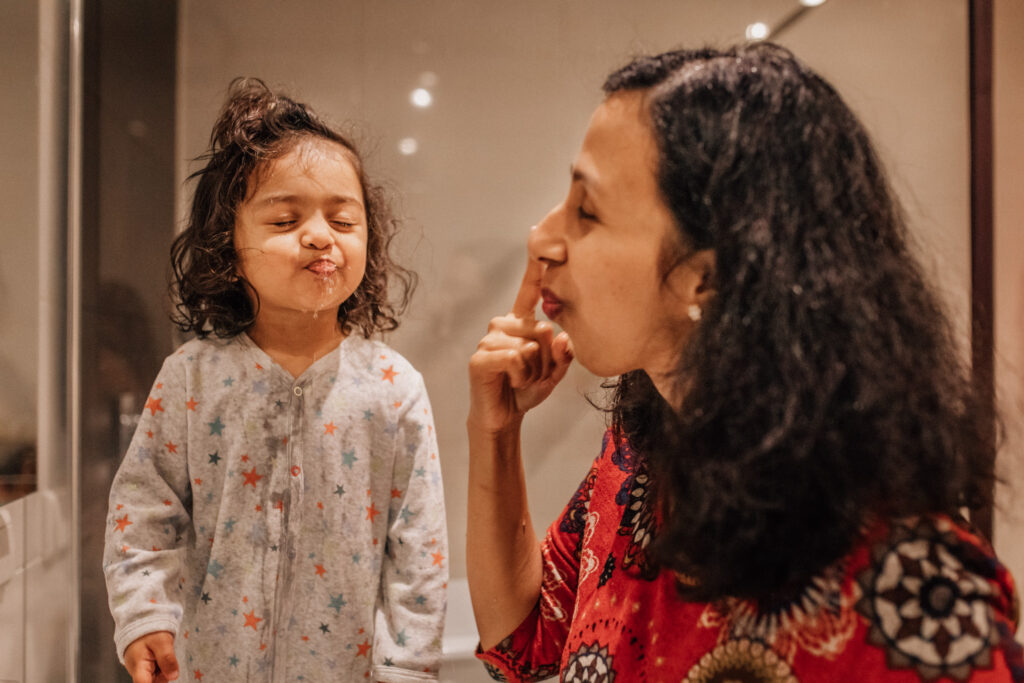 Portrait of a toddler spitting the water after washing her teeth. Her mother is next to her, showing her how to do it.