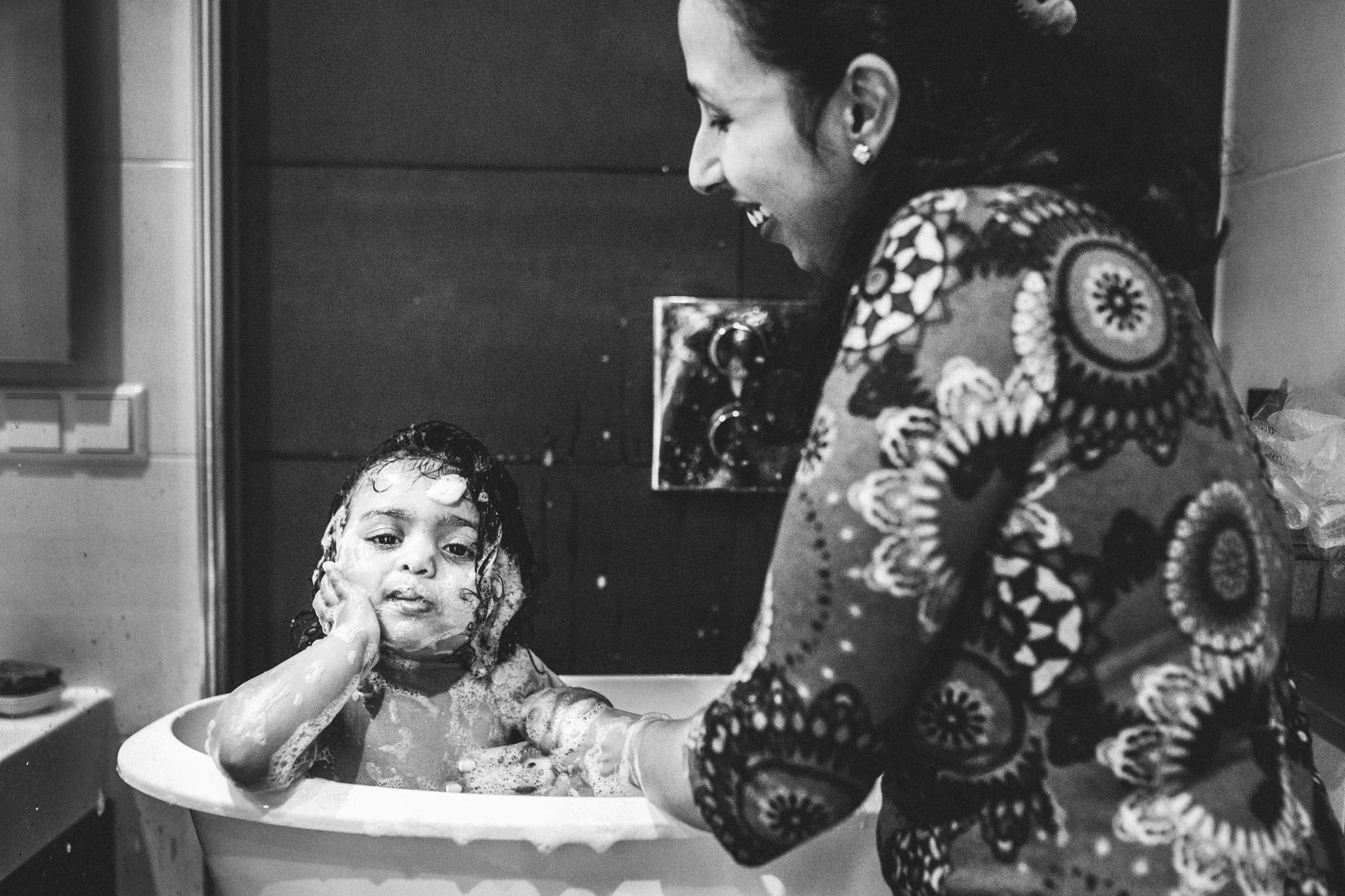 In this black and white picture, a mom is bathing her daughter. The little one is looking at the camera, her face is covered by foam. Her head is resting on her hand.