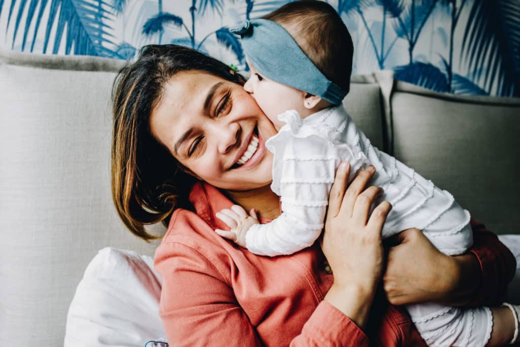 Portrait of mom playing with her little daughter. Mom has a huge smile, her hair is messy. Her baby is biting her cheek.