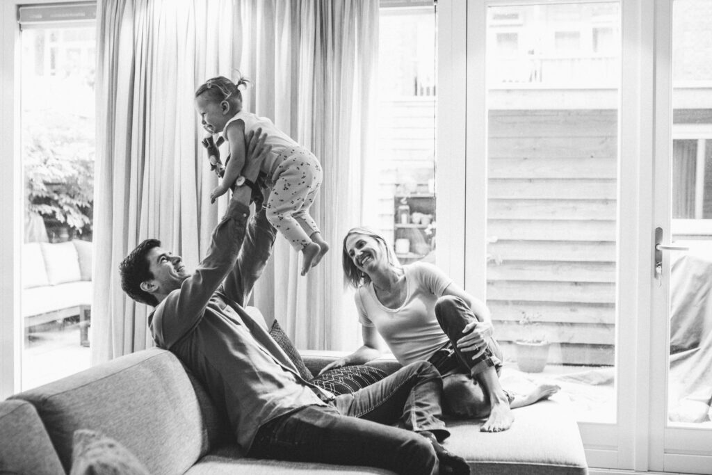 Black and white portrait of a family playing in the living room. The father is holding his daughter in the air, and the mother is smiling, looking at them.