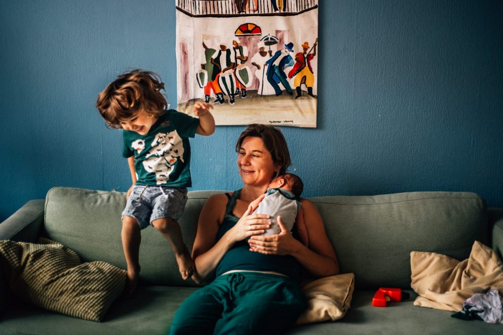 Toddler jumping on the couch where his mom is sitting holding her newborn baby. Newborn photography Amsterdam with siblings