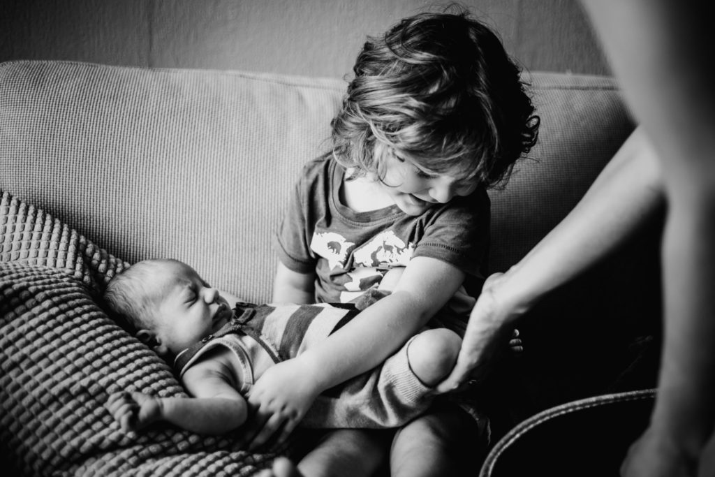 Sample of newborn photography by Lucrecia Carosi, Family and baby photographer in Amsterdam: Little boy (aprox 2 years old) holding his newborn brother for the first time. They're sitting on a couch. Moms hand is there helping him. Black and white photography. NL: Klein kind dat voor het eerst zijn pasgeboren broertje vasthoudt. Mama helpt hem daarbij. Baby fotografie.