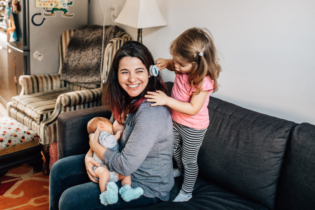 Mom is sitting on the couch breastfeeding her newborn while her older child is brushing mom's hair