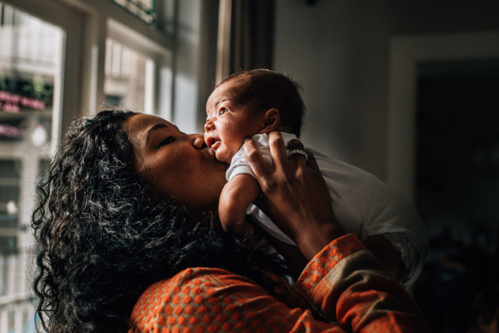 Portrait of mom holding her newborn baby, kissing his cheek. She wears a wonderful orange dress with a soft pattern, baby is wearing a white onesie.