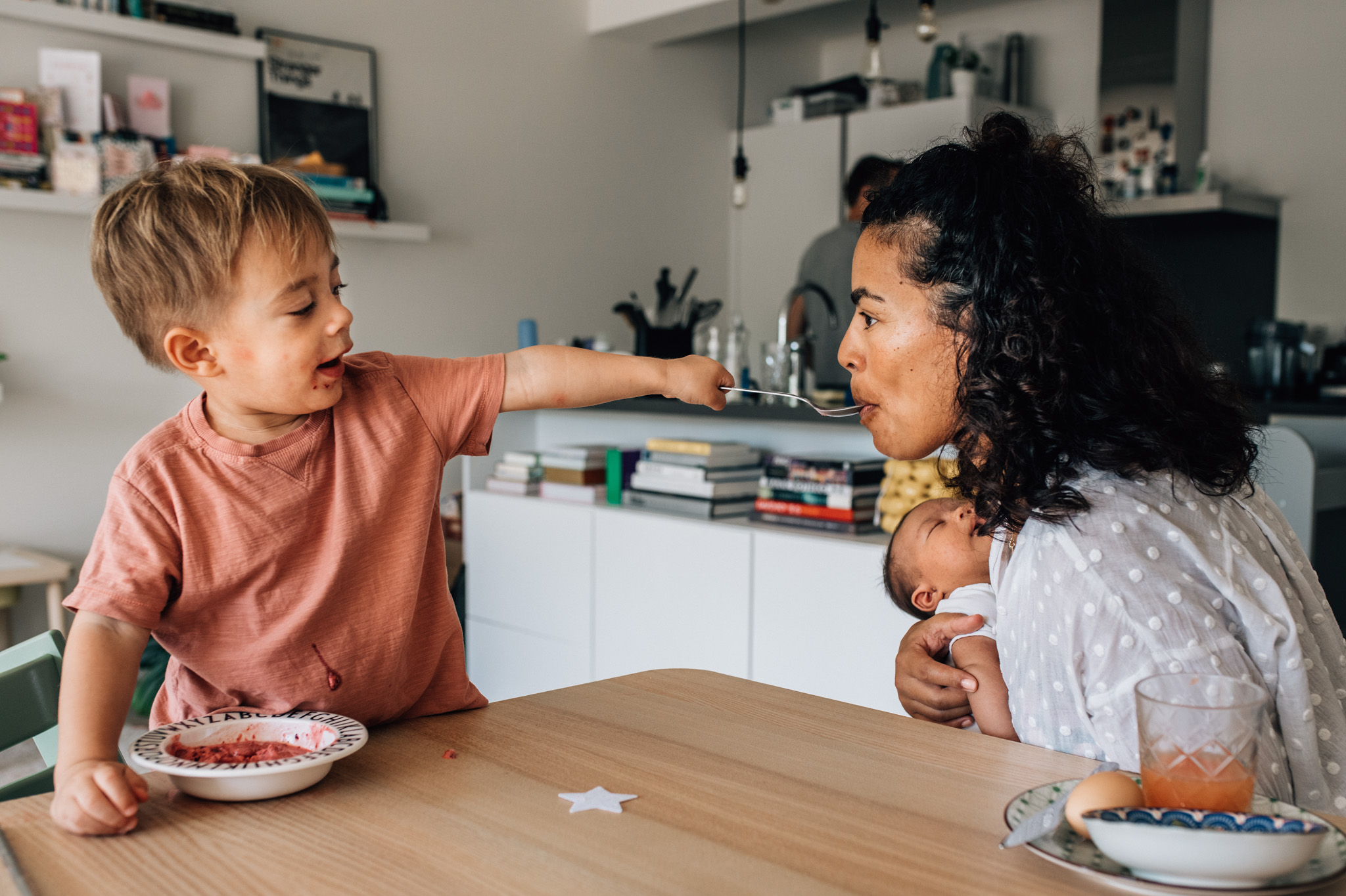 toddler feeding his mom with a spoon while she's holding her baby