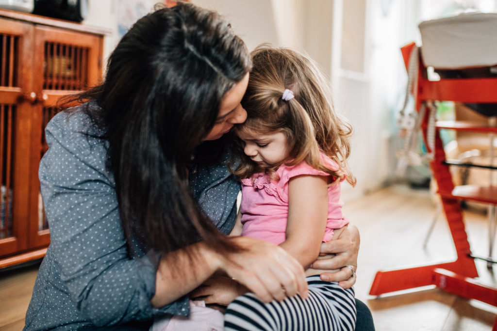 Mom kissing her toddler daughter on her front. They're sitting on the dining room floor. Mom is wearing a blue shirt and the little one is wearing a pink blouse.