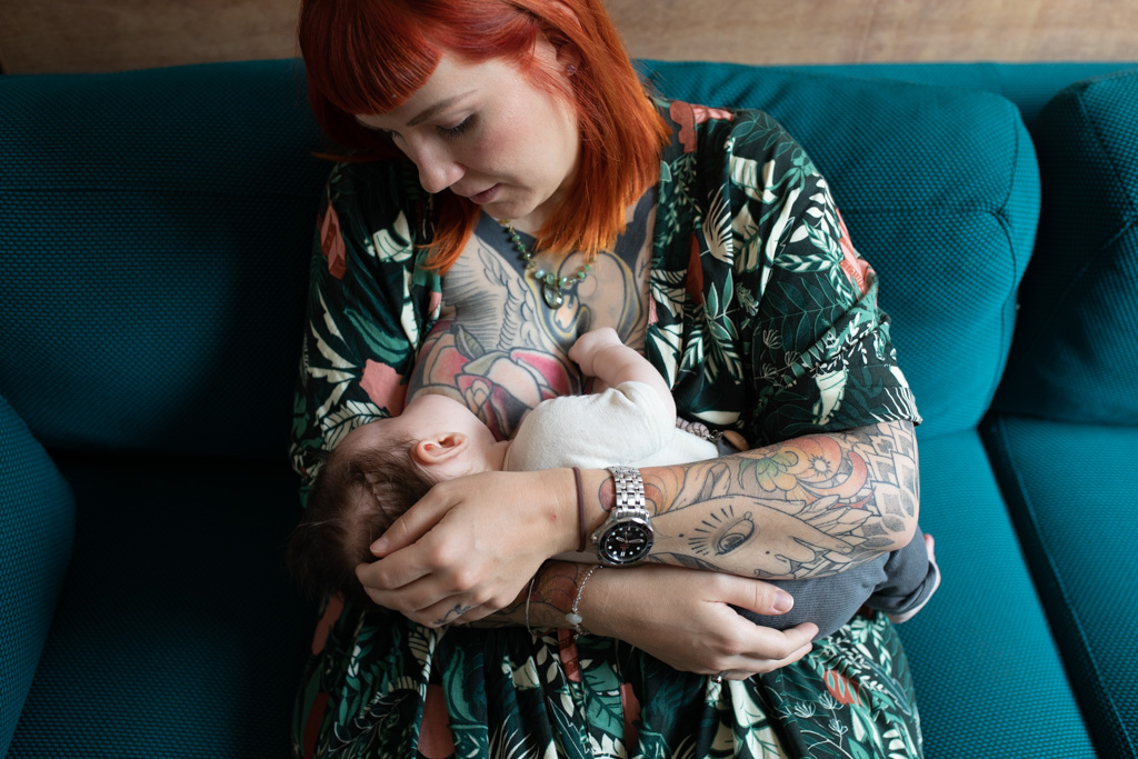 mom breastfeeding her newborn. Mom has red hair, her chest and arms are fully covered by tatooes, they're on a blue couch.