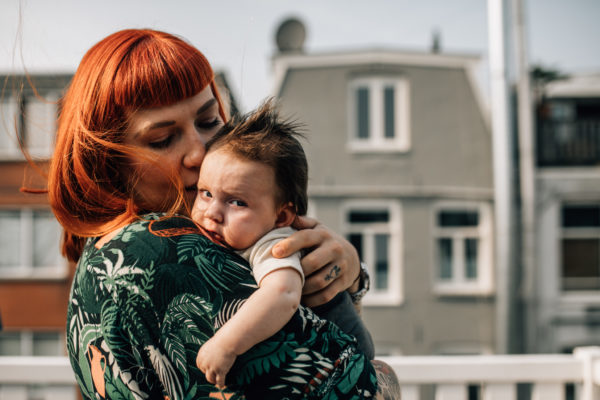 Mom holding her baby on her chest. They're on the roof of their house, Amsterdam characteristics buildings can be seen behind them. Mom has beautiful orange hair that is moving because of the wind, she has short bangs, cool tattoos on her arms and fingers, and is wearing a palm print green dress. Baby is facing the sunshine but looking at the camera, she's wearing a white onesie.