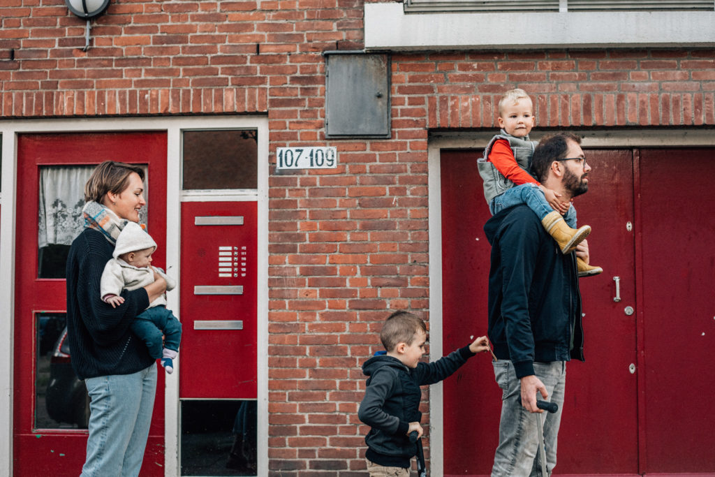 Family walking on the street, dad is on the front with one of his kids on his shoulder. Behind him, the eldest child is puting something on dad's pockets. Mom is holding the baby girl.