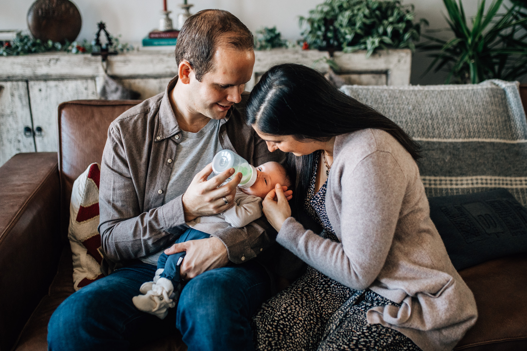 Family on the couch. Dad is feeding his newborn baby while mom is sitting next to them and caressing the little one's face.