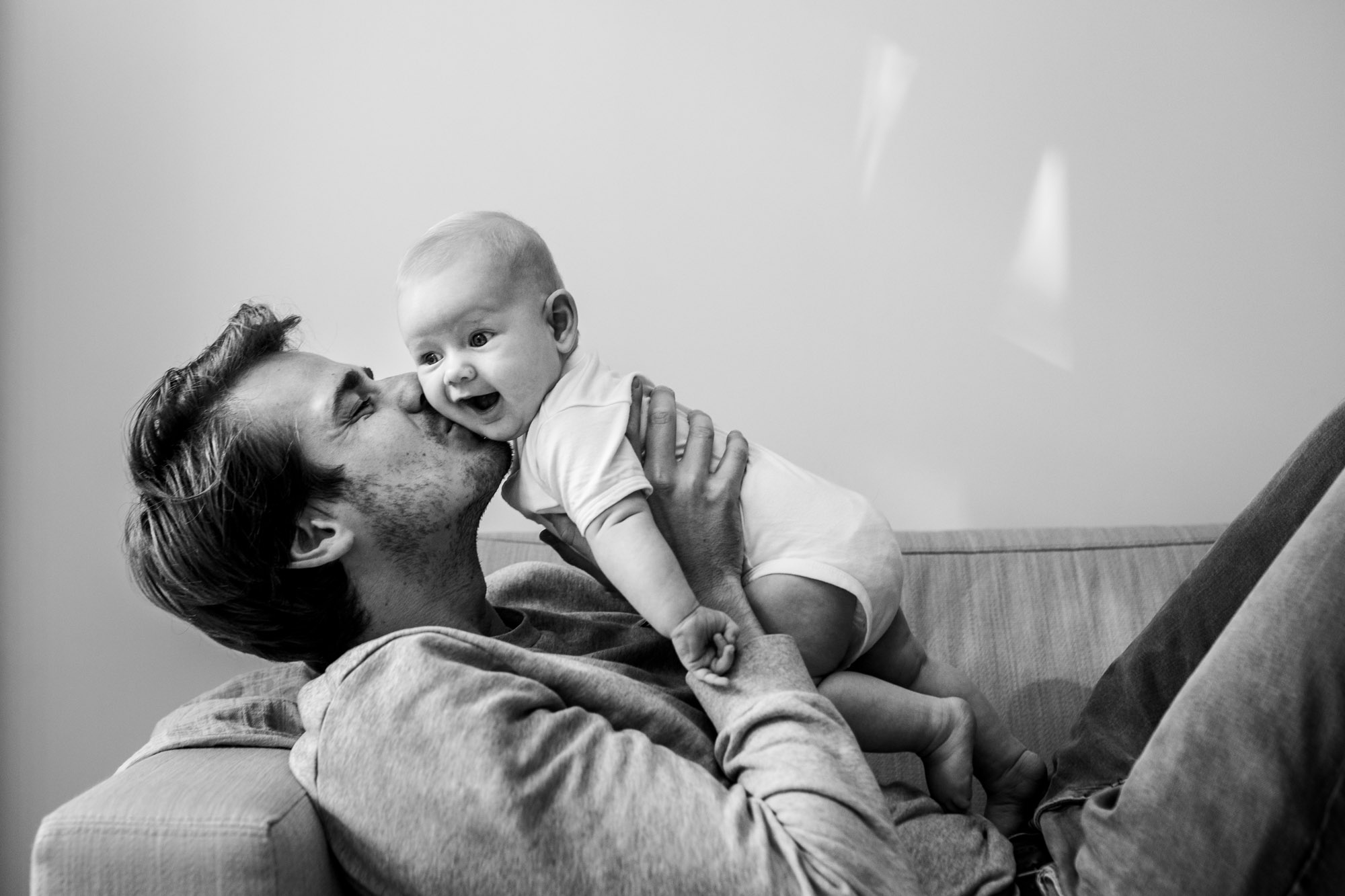 Dad kissing his baby daughter cheek while she gives a huge smile. They're on the couch. Black and white portrait.Family and baby photographer in Amsterdam
