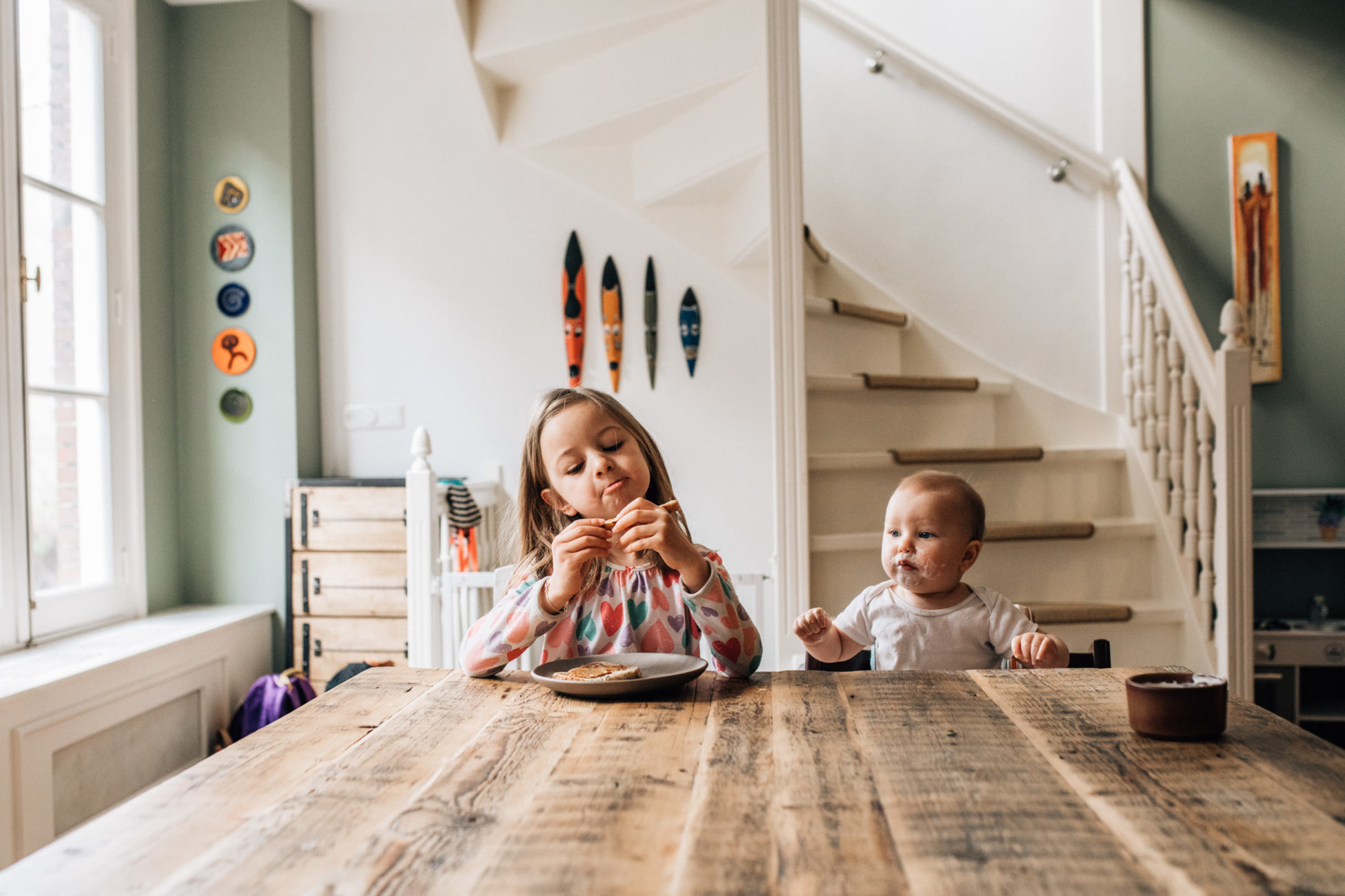 Sisters having breakfast. On the left, the older one is eating bread while the baby sitting next to her is glazing the bread very seriously.