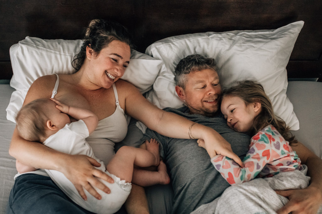 Morning cuddles. Family of 4 lying on the bed. Mom holding her baby on her chest while breastfeeding her. Next to them is dad hugging their older daughter. They're wearing pajamas.