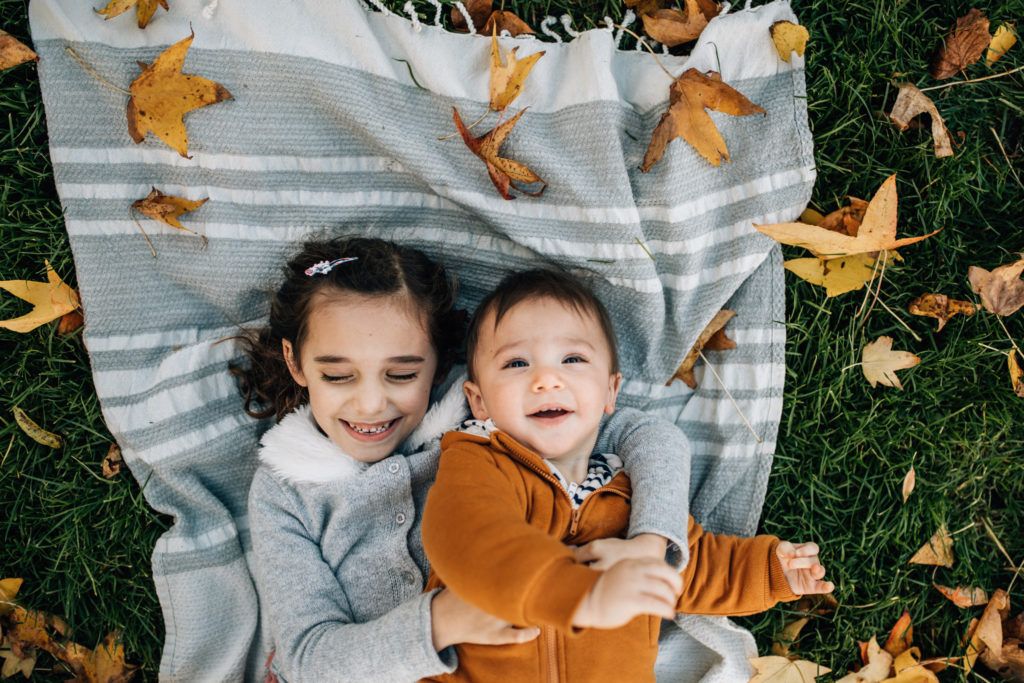 Siblings lying on a blanket in a park, there are fall leaves all around them