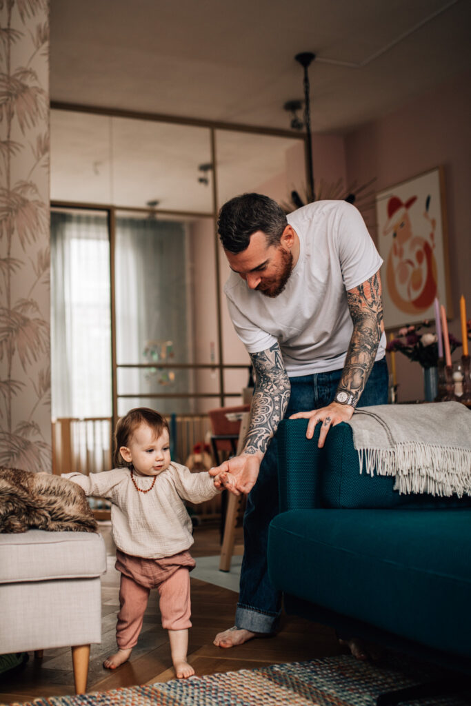 In this image, the father and the child are in their living room. The girl is trying to walk holding her dad's hand. The father's arms are fully covered in tattoos.