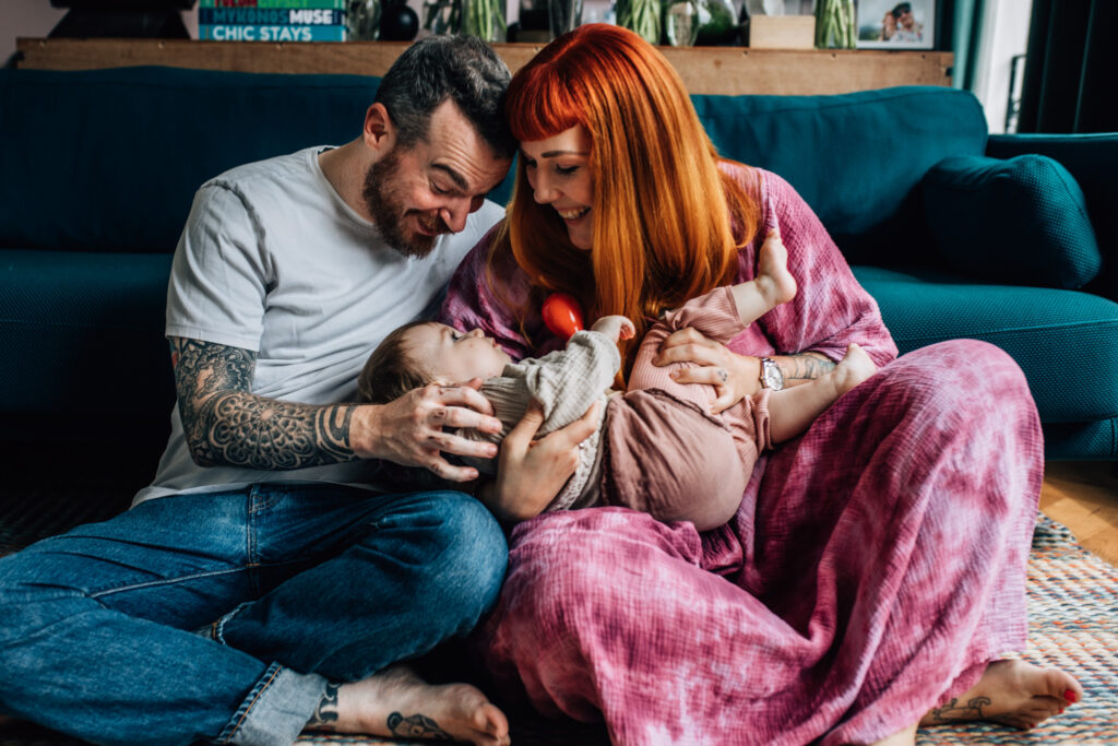 Image of an amazing family photoshoot in which both parents are holding their toddler on their lap. Mom is wearing a beautiful purple dress, dad wears jeans and a t-shirt and the toddler is wearing linnen clothes. They're sitting on the floor of their living room.