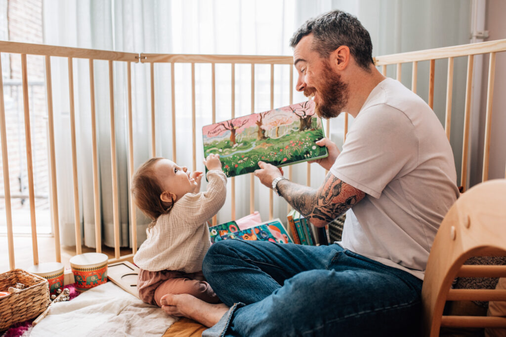 In the picture, dad is showing his toddler a book and the little one is smiling looking at him. 