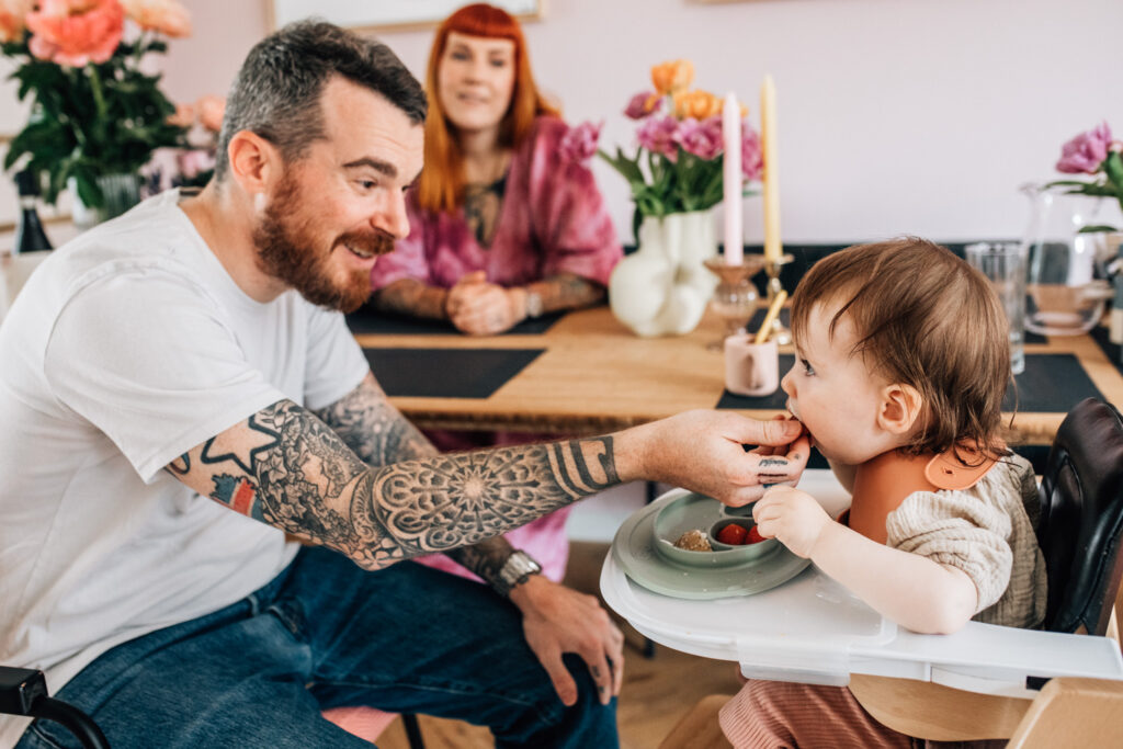 Image of a family during breakfast. The father is feeding his little toddler. She's eating fruits.