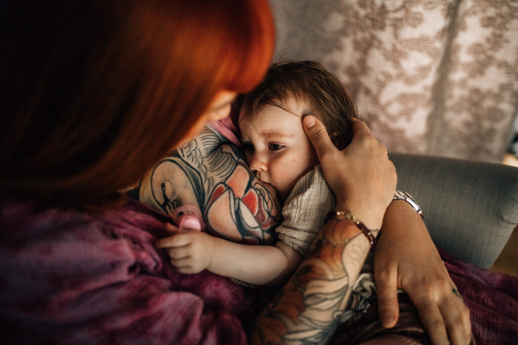 Breastfeeding image. There's very little light in the room. You can see mom is caressing her daughter, who has her eyes barely open. Mom's arms and chest are fully covered by tattoos.