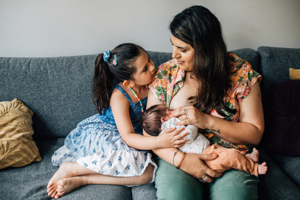 mom breastfeeding her newborn child while cuddling with her older child on the couch of their living room.