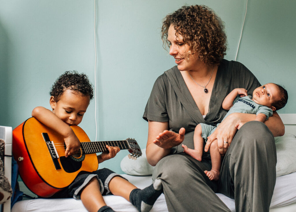 in the picture the mom is holding her newborn kid, sitting next to her toddler who is playing the guitar