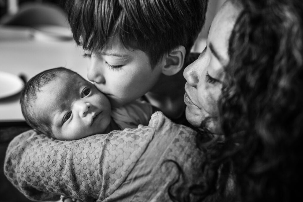 black and white portrait of mom holding her newborn while the older sibling is kissing the baby's cheek