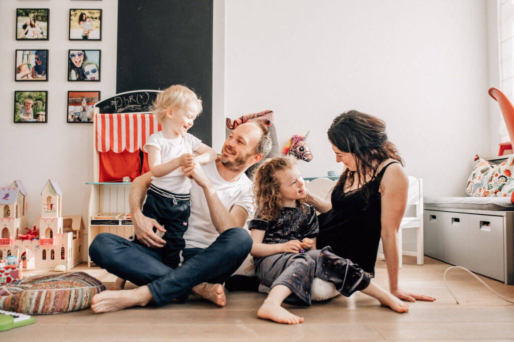 Portrait of a family sitting on the floor of the playing area in their Amsterdam's home. The father is hugging and tickling his toddler and mom is holding their daughter while caressing her hair. 