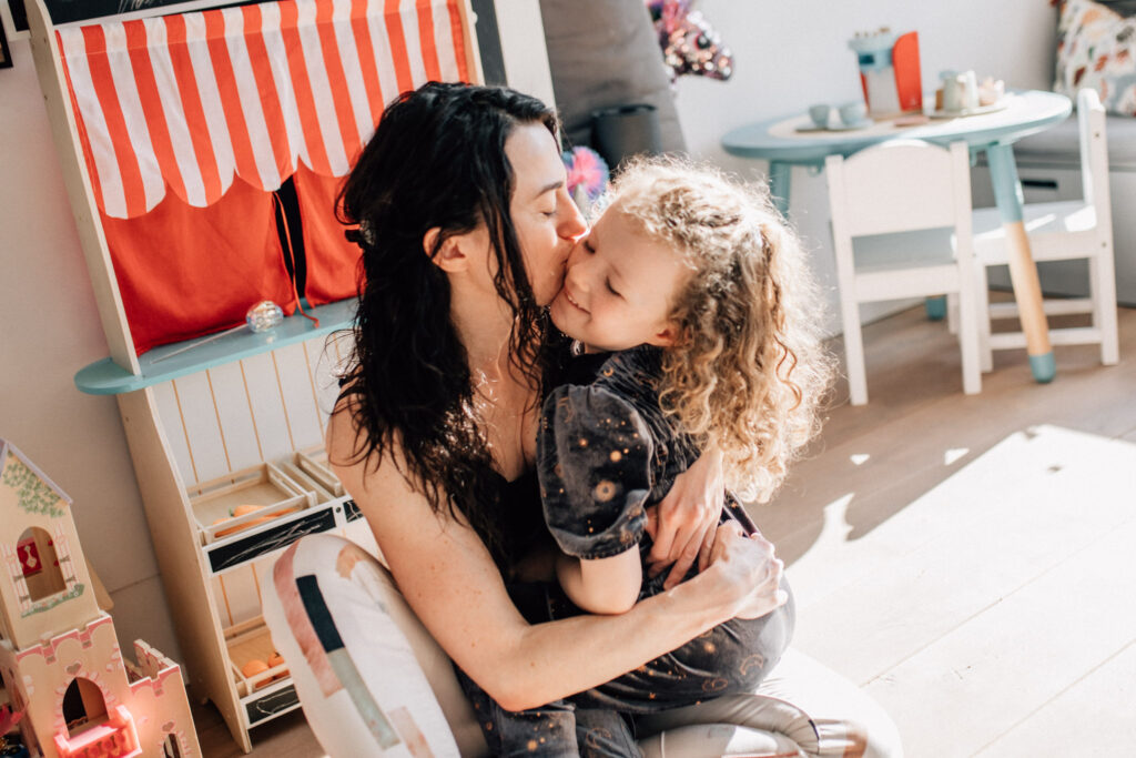 Mother hugging and kissing her little daughter in an area of their house that is full of toys. The picture belong to a family photography session in Amsterdam.