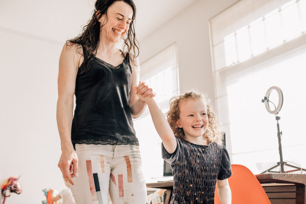 Photography of a girl smiling, walking in her house, holding her mom's hands. Mom is also smiling. In-home family photography allows children to feel confident and safe, and getting beautiful genuine smiles from them. 